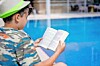 Child wearing a green cap reads an open book beside a blue swimming pool.