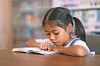 A young child reads an open book at a table with bookshelves behind.