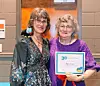 Two women standing indoors, one holding a framed 30 years of service certificate.