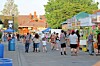Crowd walking past market stalls and tents on a street with buildings and trees in the background.