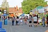 Crowd walking past market stalls and tents on a street with buildings and trees in the background.