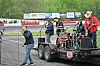 A band performs on a trailer stage with drums, microphones and amplifiers beside an outdoor track.