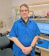 Man in a blue shirt standing in an office beside a desk with computer monitors.