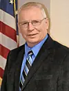 A man wearing glasses, a dark suit, and a striped tie poses in front of an American flag.