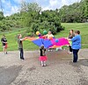 Children and adults hold a rainbow parachute with balls on top in a park.
