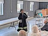 A woman addresses a seated audience inside a hall with tables and a rolling cart behind her.