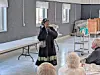 A woman addresses a seated audience inside a hall with tables and a rolling cart behind her.