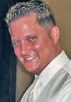 Smiling man in a white shirt, tie, and light vest against a dark background.