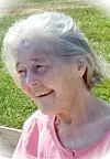 Elderly woman smiling outdoors beside a grassy area.