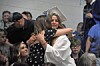 A graduate in a white cap and gown hugs a woman in a black dress in a crowded ceremony.