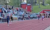 Runners race on a track beside bleachers filled with spectators at an outdoor stadium.