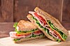 Two stacked sandwich halves with deli meat, lettuce and tomato on a wooden cutting board.