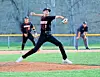 Baseball pitcher in black uniform throwing a pitch on a field, with two people behind him.