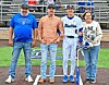 Four people standing on a baseball field near home plate with stadium bleachers behind them.