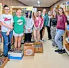 Students standing in a school hallway next to boxes filled with donated food and supplies.