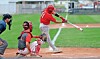Batter in red swings at a pitch with catcher and umpire behind home plate.