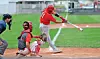 Batter in red swings at a pitch with catcher and umpire behind home plate.