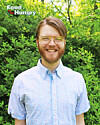 Smiling man with glasses in a light blue shirt standing before green foliage.
