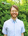 Smiling man with glasses in a light blue shirt standing before green foliage.