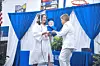 Woman holding a child with a man in white at a ribbon-cutting setup with blue drapes and balloons.