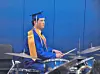 Graduate in blue cap and gown seated at a drum kit, holding drumsticks.