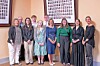 Eight people stand in a line indoors in front of framed photo collages.