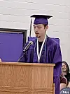 A graduate in purple cap and gown stands behind a wooden podium and microphone, speaking indoors.