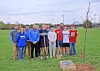 A group of people standing on grass beside a newly planted tree in an open field.