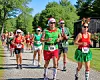 Runners in Santa hats and holiday costumes compete on a paved road surrounded by trees.