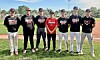 Seven baseball players stand side by side on grass in front of a baseball diamond.