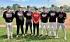 Seven baseball players stand side by side on grass in front of a baseball diamond.