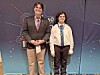 Two people wearing medals stand on a stage in front of an event backdrop.