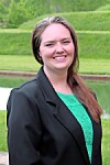 Smiling woman in a black blazer and green top stands outdoors in front of green hedges.