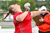 Athlete in red throws a shot put outdoors while an official stands nearby with a clipboard.