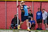 Athlete in blue and black uniform throws in a track and field cage with spectators behind a net.