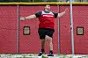 Track and field athlete in red and black gear preparing to throw in front of a red wall.
