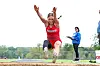 Athlete in red lands in the sand during a long jump at an outdoor track and field facility.