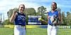 Two softball players pose on a field with a scoreboard in the background.