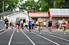 Several runners race down an outdoor track with lane markings, trees, buildings and solar panels behind them.