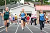 Three runners race on a track with other people and tents in the background.