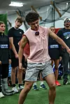 Teenage athlete in a pink sleeveless shirt and patterned shorts practices indoors with others standing behind him.