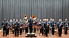 A large orchestra stands on a concert hall stage with instruments and music stands arranged in front.