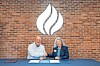 Two people seated at a table in front of a brick wall with a large white logo above.