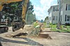 Construction workers and an excavator dig a trench on a street near apartment buildings.