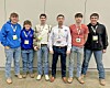 Six people standing side by side indoors, facing the camera and wearing event badges.