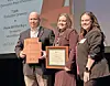 Three people on stage holding a framed certificate and an award plaque at a recognition ceremony.