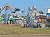 People near colorful amusement rides and fenced attractions at a fairground.