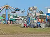 People near colorful amusement rides and fenced attractions at a fairground.