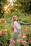 Smiling woman stands among pink and orange flowers in a sunlit garden.