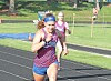 Runner in blue and red uniform leading on an outdoor track with competitors behind.